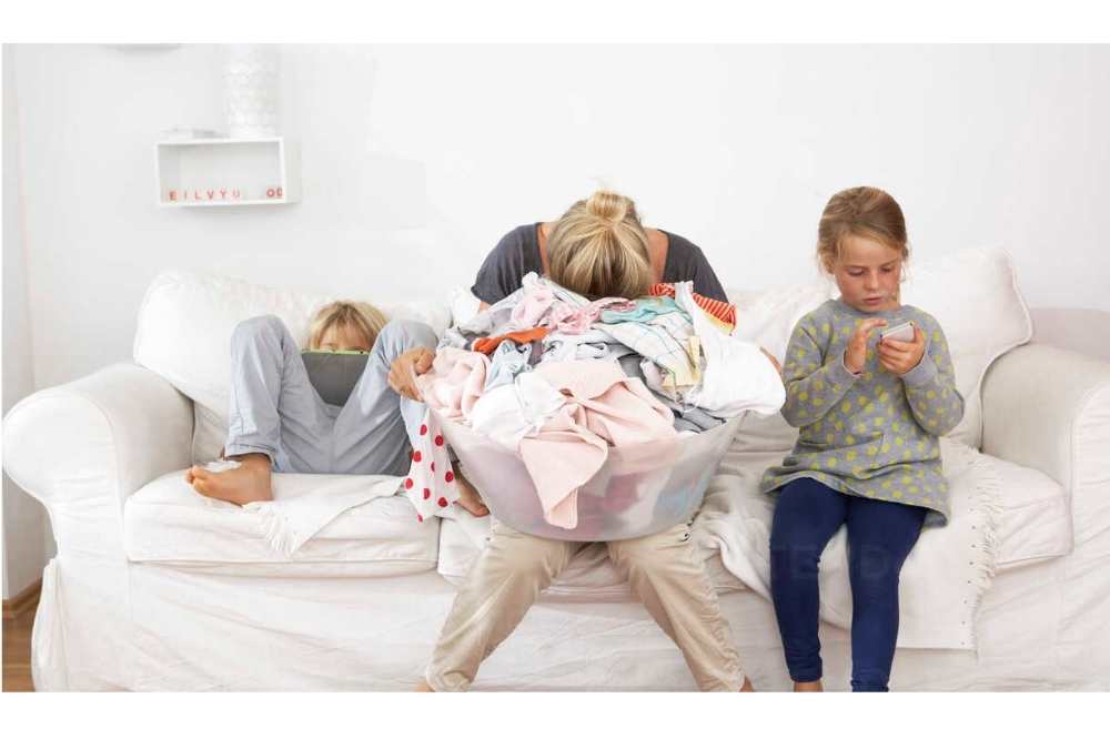 Eine Mutter mit übervollem Wäschekorb sitzt erschöpft zwischen zwei Kindern, die am Tablet und Handy spielen – – Sinnbild für Mom Rage und stille Überforderung im Familienalltag